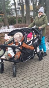 Opiekunka żłobka prowadząca wieloosobowy wózek spacerowy z dziećmi podczas zimowego spaceru na terenie placówki