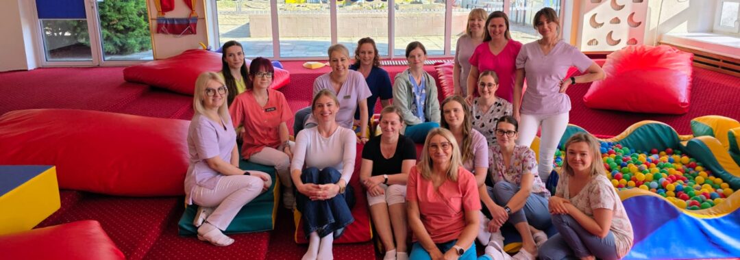 Zespół opiekunów żłobka podczas szkolenia prowadzonego przez fizjoterapeutkę – wspólne zdjęcie uczestników w sali zabaw.
