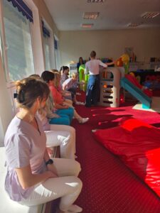 Część praktyczna szkolenia – fizjoterapeutka pokazuje opiekunom prawidłowe ułożenie ciała i postawę podczas pracy z dzieckiem.