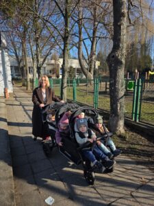 Opiekunki ze Żłobka Pomnik Matki Polki prowadzą wózki wieloosobowe z dziećmi podczas spaceru chodnikiem w słoneczny dzień.