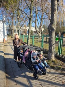 Opiekunki ze Żłobka Pomnik Matki Polki prowadzą wózki wieloosobowe z dziećmi podczas spaceru chodnikiem w słoneczny dzień.
