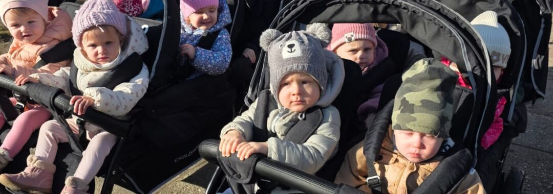 Opiekunki ze Żłobka Pomnik Matki Polki prowadzą wózki wieloosobowe z dziećmi podczas spaceru chodnikiem w słoneczny dzień.