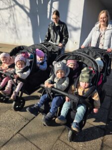 Opiekunki ze Żłobka Pomnik Matki Polki prowadzą wózki wieloosobowe z dziećmi podczas spaceru chodnikiem w słoneczny dzień.