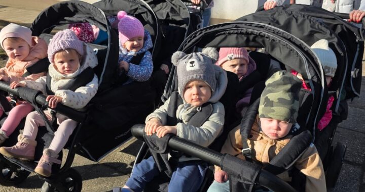 Opiekunki ze Żłobka Pomnik Matki Polki prowadzą wózki wieloosobowe z dziećmi podczas spaceru chodnikiem w słoneczny dzień.