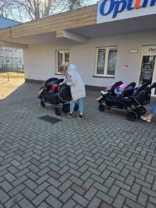 Opiekunki ze Żłobka Pomnik Matki Polki prowadzą wózki wieloosobowe z dziećmi podczas spaceru chodnikiem w słoneczny dzień.