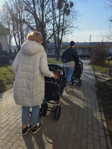 Opiekunki ze Żłobka Pomnik Matki Polki prowadzą wózki wieloosobowe z dziećmi podczas spaceru chodnikiem w słoneczny dzień.