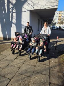 Opiekunki ze Żłobka Pomnik Matki Polki prowadzą wózki wieloosobowe z dziećmi podczas spaceru chodnikiem w słoneczny dzień.