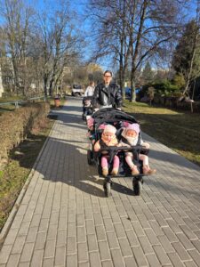Opiekunki ze Żłobka Pomnik Matki Polki prowadzą wózki wieloosobowe z dziećmi podczas spaceru chodnikiem w słoneczny dzień.