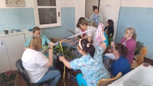 Pracownicy żłobka wspólnie przygotowują palmy wielkanocne – siedzą przy stole i układają gałązki bazi, bukszpanu oraz kolorowe wstążki. Na zdjęciach widoczne są etapy tworzenia palm oraz gotowe, bogato zdobione kompozycje