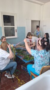 Pracownicy żłobka wspólnie przygotowują palmy wielkanocne – siedzą przy stole i układają gałązki bazi, bukszpanu oraz kolorowe wstążki. Na zdjęciach widoczne są etapy tworzenia palm oraz gotowe, bogato zdobione kompozycje