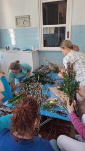 Pracownicy żłobka wspólnie przygotowują palmy wielkanocne – siedzą przy stole i układają gałązki bazi, bukszpanu oraz kolorowe wstążki. Na zdjęciach widoczne są etapy tworzenia palm oraz gotowe, bogato zdobione kompozycje