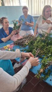 Pracownicy żłobka wspólnie przygotowują palmy wielkanocne – siedzą przy stole i układają gałązki bazi, bukszpanu oraz kolorowe wstążki. Na zdjęciach widoczne są etapy tworzenia palm oraz gotowe, bogato zdobione kompozycje