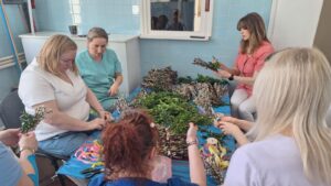 Pracownicy żłobka wspólnie przygotowują palmy wielkanocne – siedzą przy stole i układają gałązki bazi, bukszpanu oraz kolorowe wstążki. Na zdjęciach widoczne są etapy tworzenia palm oraz gotowe, bogato zdobione kompozycje