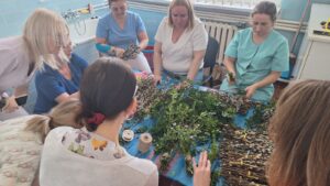 Pracownicy żłobka wspólnie przygotowują palmy wielkanocne – siedzą przy stole i układają gałązki bazi, bukszpanu oraz kolorowe wstążki. Na zdjęciach widoczne są etapy tworzenia palm oraz gotowe, bogato zdobione kompozycje