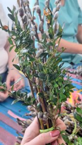 Pracownicy żłobka wspólnie przygotowują palmy wielkanocne – siedzą przy stole i układają gałązki bazi, bukszpanu oraz kolorowe wstążki. Na zdjęciach widoczne są etapy tworzenia palm oraz gotowe, bogato zdobione kompozycje