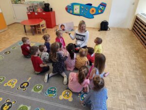 Dzieci siedząc na dywanie słuchają książki czytanej przez panią z biblioteki