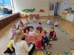 Dzieci siedząc na dywanie słuchają książki czytanej przez panią z biblioteki