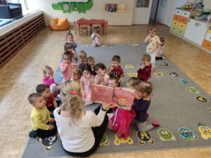 Dzieci siedząc na dywanie słuchają książki czytanej przez panią z biblioteki