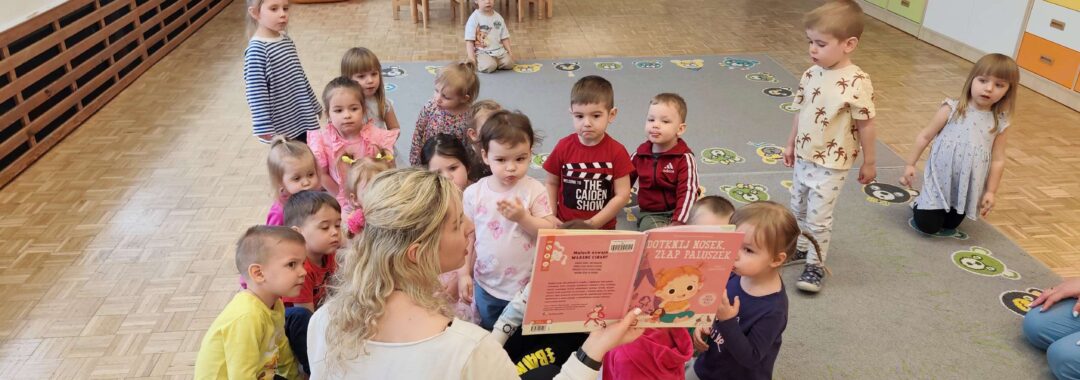 Dzieci siedząc na dywanie słuchają książki czytanej przez panią z biblioteki