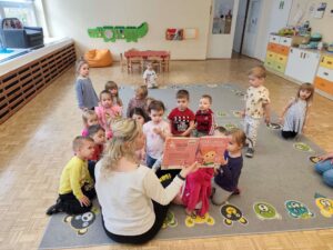 Dzieci siedząc na dywanie słuchają książki czytanej przez panią z biblioteki