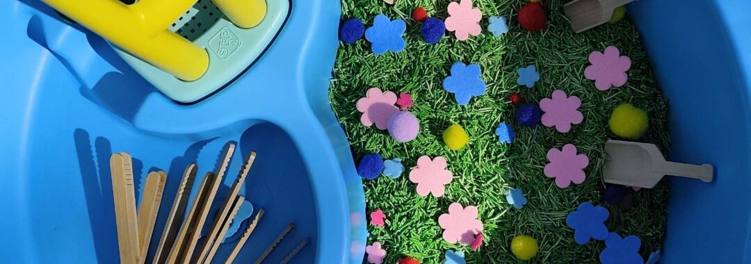 Basenik sensoryczny wypełnionym barwionym na zielono makaronem, filcowymi kwiatkami i kolorowymi pomponami, drewnianymi pęsetami i łopatkami