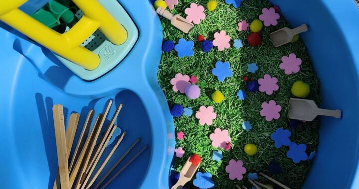 Basenik sensoryczny wypełnionym barwionym na zielono makaronem, filcowymi kwiatkami i kolorowymi pomponami, drewnianymi pęsetami i łopatkami