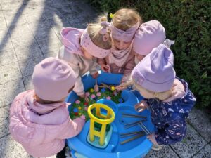 Dzieci bawią się przy baseniku sensorycznym wypełnionym barwionym na zielono makaronem, filcowymi kwiatkami i kolorowymi pomponami
