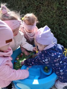 Dzieci bawią się przy baseniku sensorycznym wypełnionym barwionym na zielono makaronem, filcowymi kwiatkami i kolorowymi pomponami
