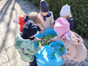 Dzieci bawią się przy baseniku sensorycznym wypełnionym barwionym na zielono makaronem, filcowymi kwiatkami i kolorowymi pomponami