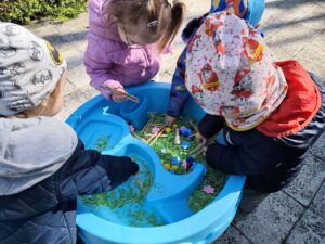 Dzieci bawią się przy baseniku sensorycznym wypełnionym barwionym na zielono makaronem, filcowymi kwiatkami i kolorowymi pomponami