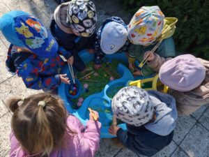 Dzieci bawią się przy baseniku sensorycznym wypełnionym barwionym na zielono makaronem, filcowymi kwiatkami i kolorowymi pomponami